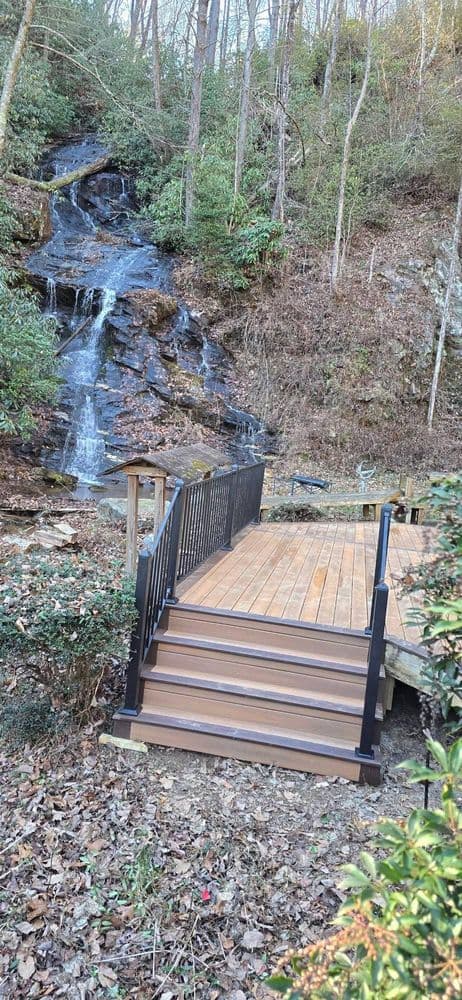Wooden deck with black railing beside a cascading waterfall in a wooded area.