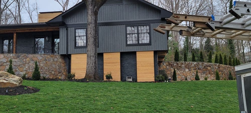 Modern house with stone walls, large windows, and greenery in the front yard.