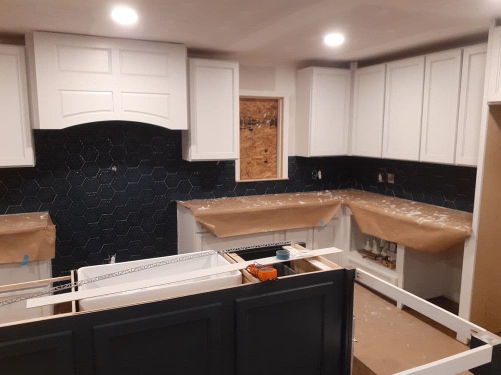 Renovated kitchen with white cabinets, dark hexagon backsplash, and unfinished cabinetry.