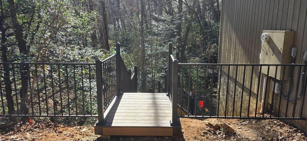 Wooden deck with metal railing leading into a wooded area, surrounded by trees.