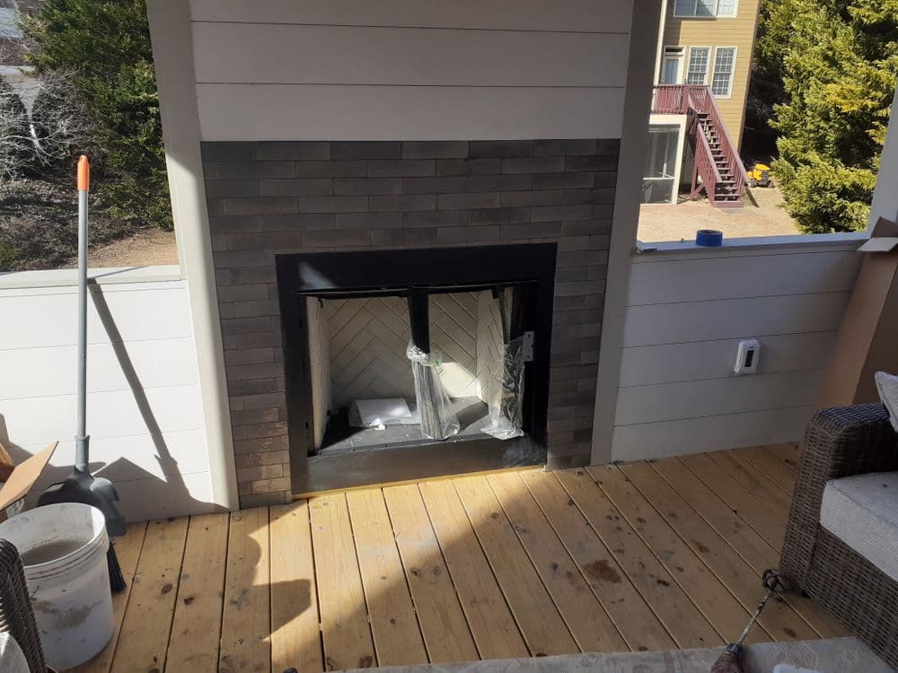 Modern outdoor fireplace with a stone surround on a wooden patio, surrounded by greenery.