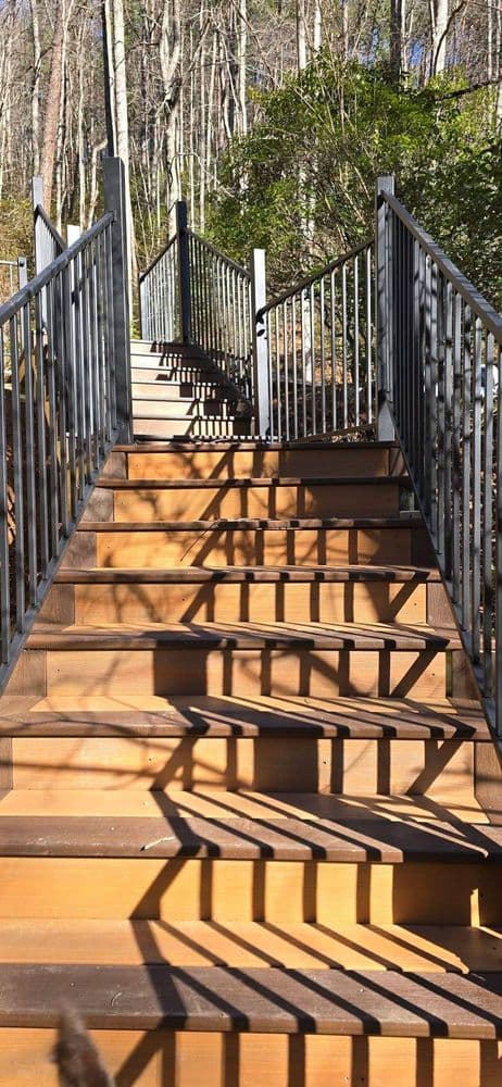 Wooden staircase with metal railing in a wooded area, sunlight casting shadows.