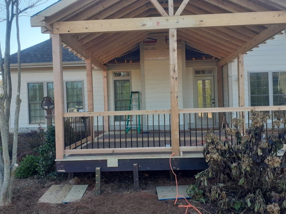 Newly constructed front porch with wooden framework and railing, surrounded by landscaping.
