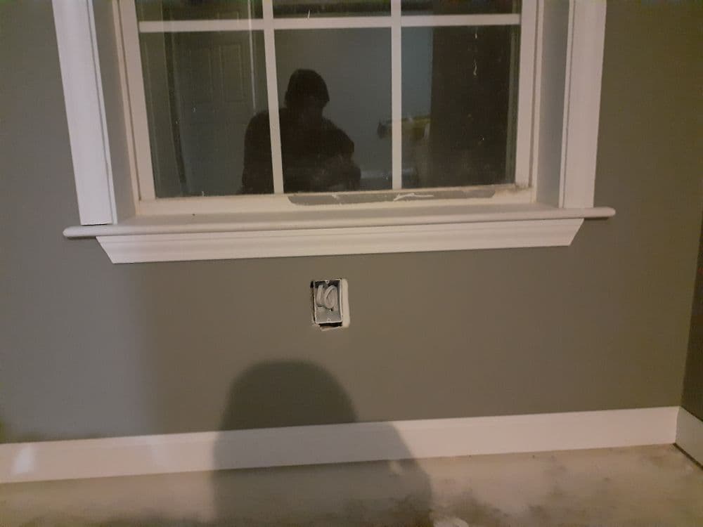 Interior wall with window and electrical outlet during renovation, showing gray paint and trim.