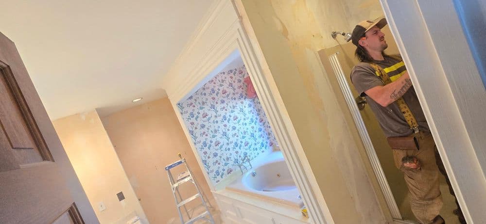 Man renovating a bathroom with floral wallpaper and a bathtub in the background.