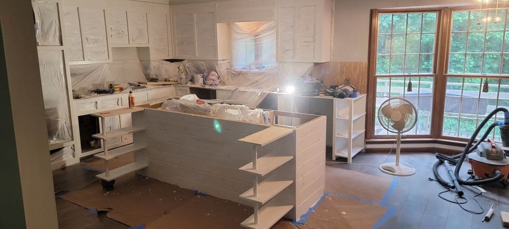 Renovated kitchen with white cabinets, unfinished island, construction materials, and natural light.