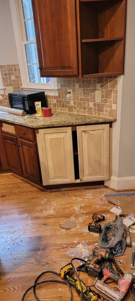 Messy kitchen renovation with unfinished cabinets, tools, and debris on wooden floor.
