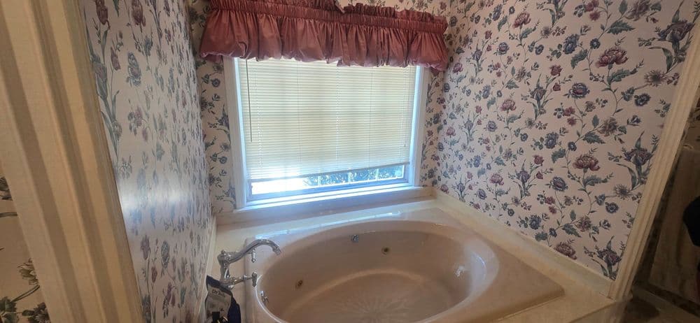 Cozy bathroom featuring a jetted tub, floral wallpaper, and a window with blinds.