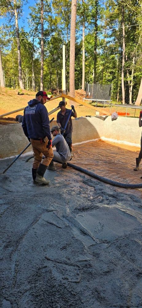 Workers pouring concrete for a swimming pool in a forested outdoor setting.