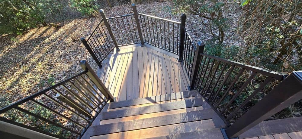 Wooden deck with black railings surrounded by trees and fallen leaves, viewed from above.