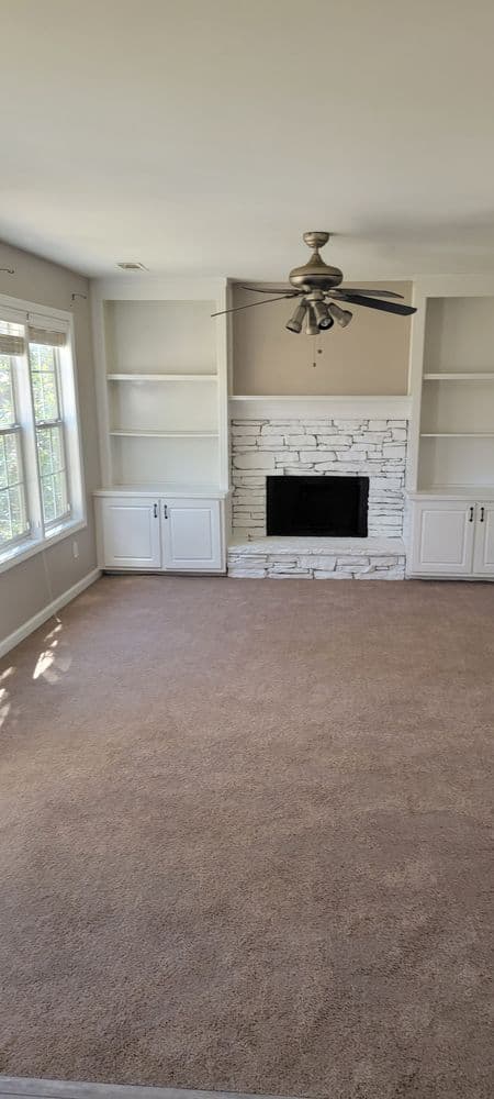 Cozy living room with light carpet, stone fireplace, and ceiling fan, featuring built-in shelves.